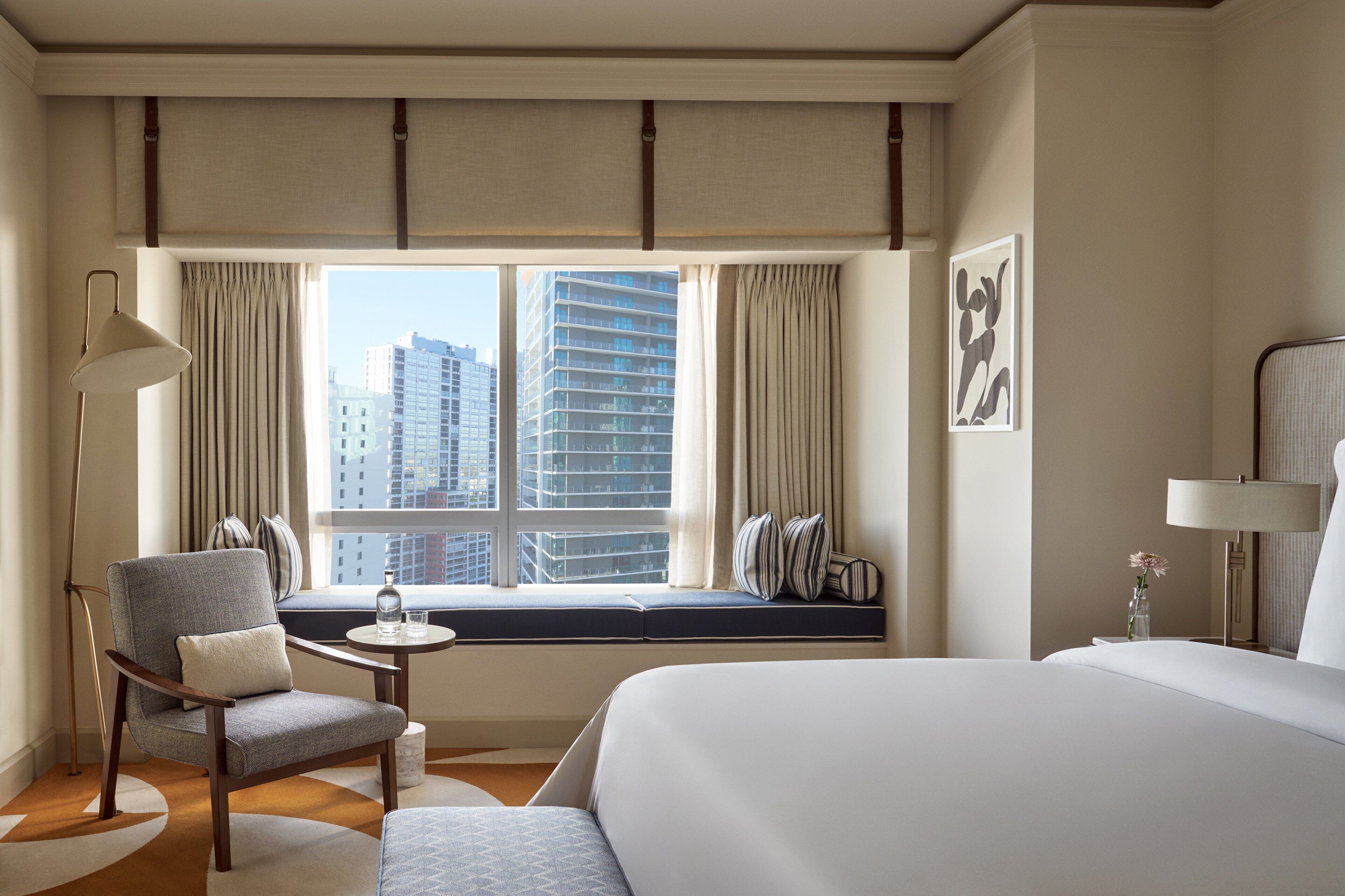 Een hotelkamer met een groot bed, een zithoek met een stoel en een klein tafeltje, een raam met gordijnen en uitzicht op de stad.
