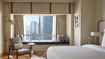 Een hotelkamer met een groot bed, een zithoek met een stoel en een klein tafeltje, een raam met gordijnen en uitzicht op de stad.