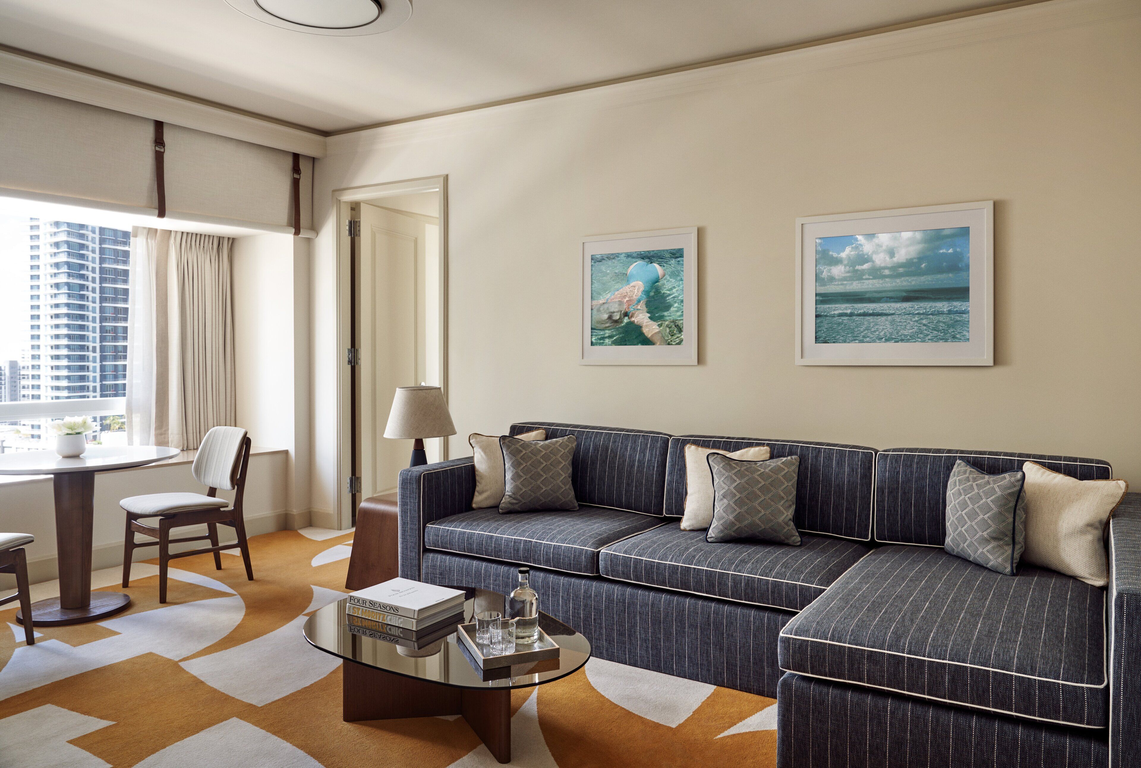 A modern living room with a sectional sofa, a glass coffee table, and a dining area with a table and chairs. There are two framed pictures on the wall.
