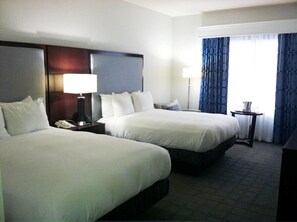 A hotel room with two beds, a nightstand with a lamp, a small table with a pitcher, and a window with blue curtains.