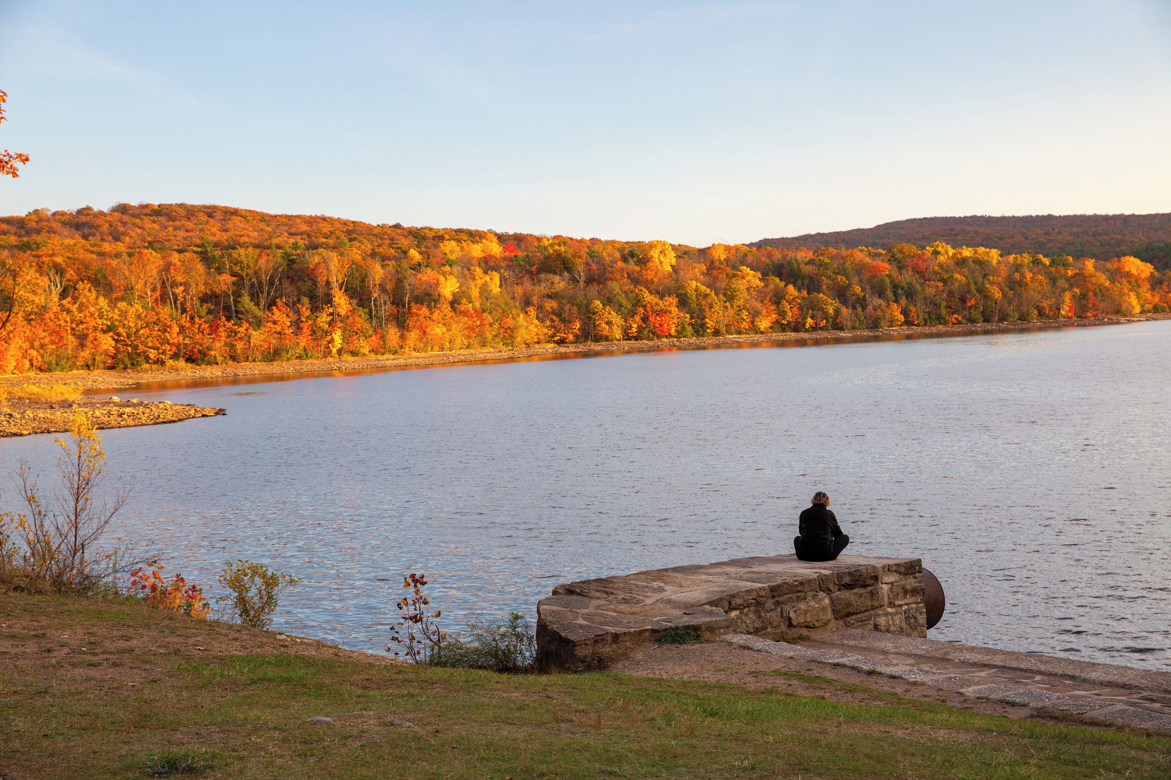 A person sitting on a rock by a calm lake with autumn foliage in the background.