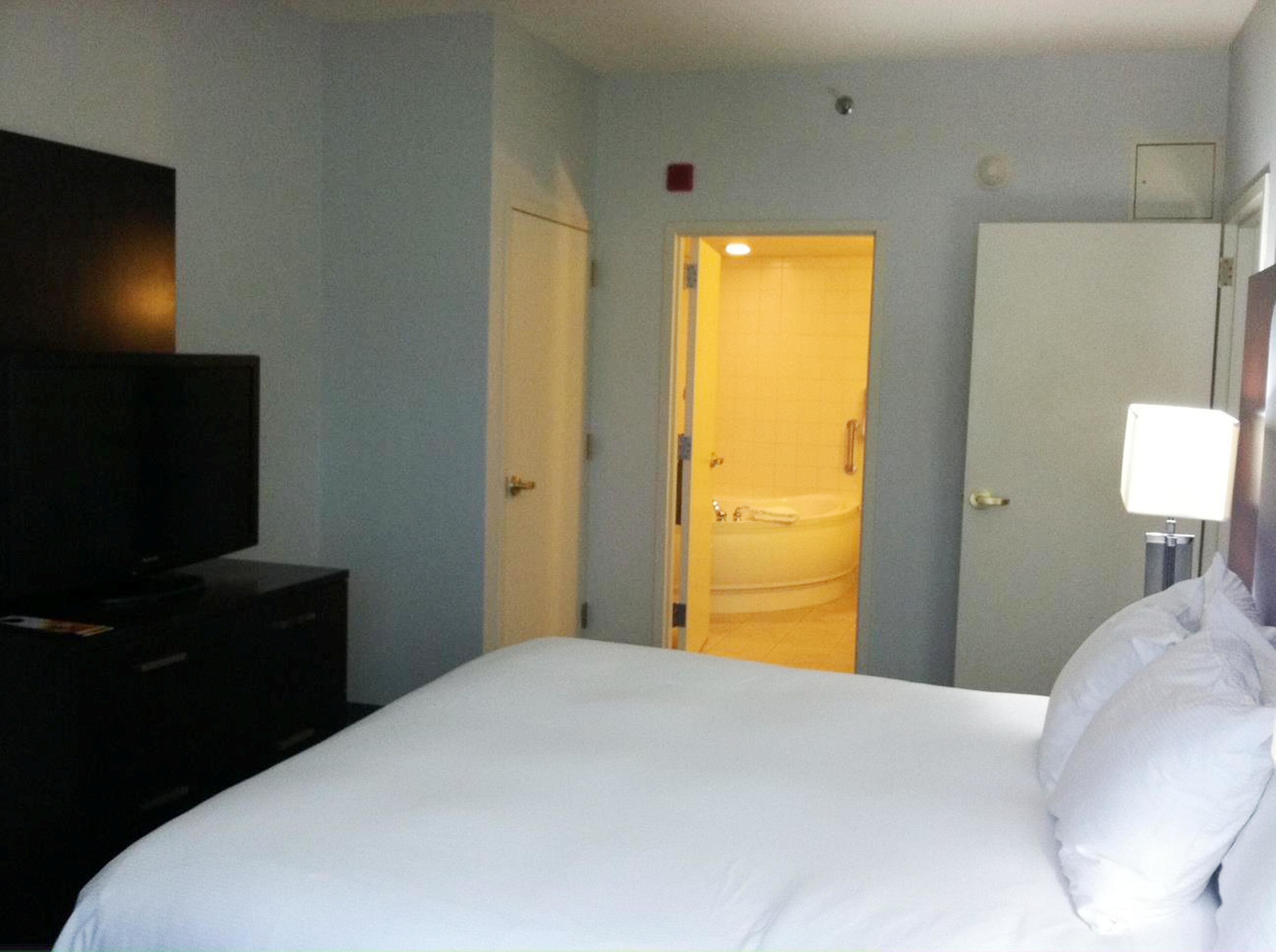 A hotel room with a bed, a TV on a dresser, a bathroom with a sink and bathtub, and a door leading to another room.