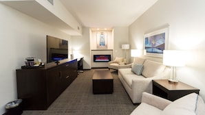 A modern hotel room with a flat-screen TV, a dark wooden entertainment unit, a beige sofa, a coffee table, and a wall-mounted fireplace. - Hilton Scranton & Conference Center (Scranton)