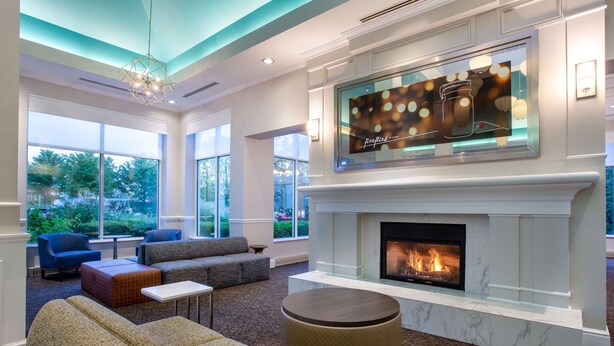 A spacious living room with a fireplace, large windows, and a modern chandelier. - Hilton Garden Inn Islip/MacArthur Airport (Ronkonkoma)