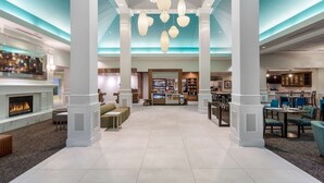 A spacious lobby with a central fireplace, a bar area, and a seating area with tables and chairs. - Hilton Garden Inn Islip/MacArthur Airport (Ronkonkoma)