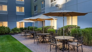 An outdoor patio area with brick flooring, round tables, chairs, and large umbrellas. - Hilton Garden Inn Islip/MacArthur Airport (Ronkonkoma)
