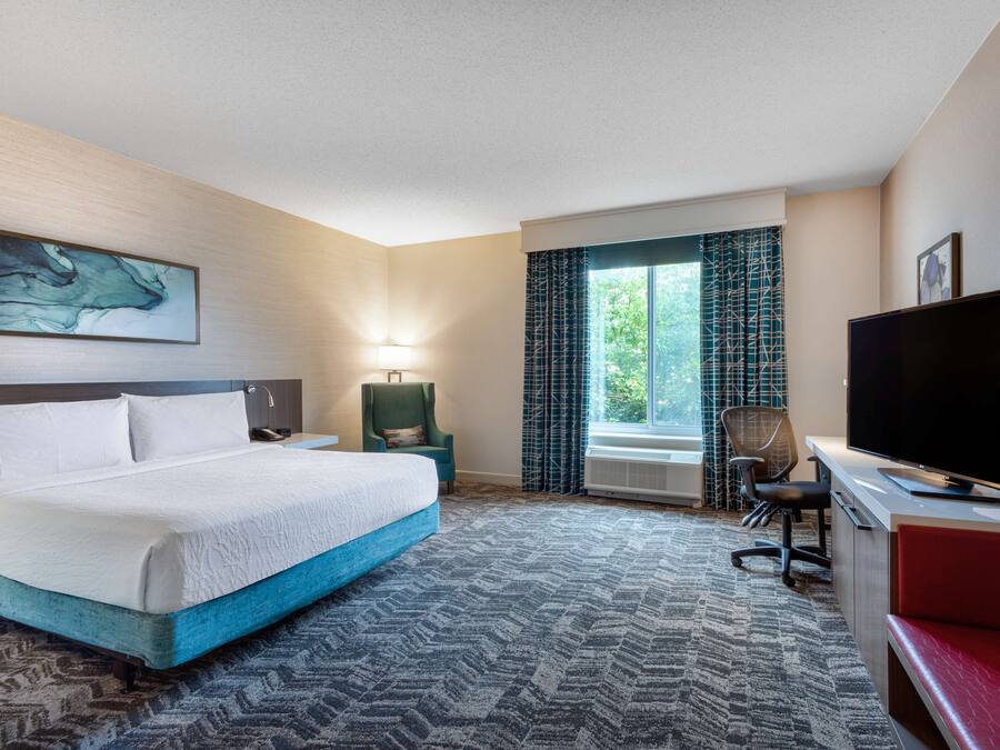 A hotel room with a bed, a desk with a TV, a chair, a lamp, and a window with curtains.