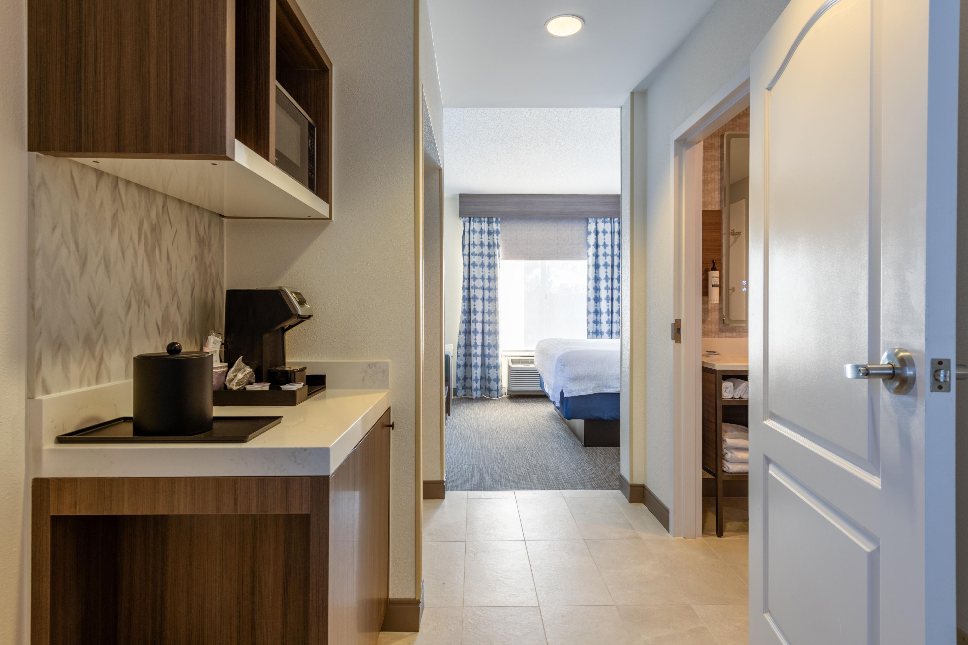 A hotel hallway with a kitchen area on the left, a bed with blue curtains in the center, and a bathroom visible through an open door on the right.