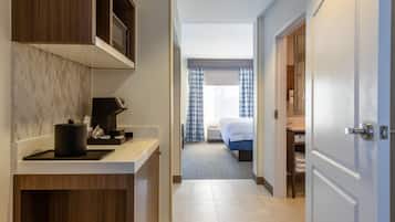 A hotel hallway with a kitchen area on the left, a bed with blue curtains in the center, and a bathroom visible through an open door on the right.