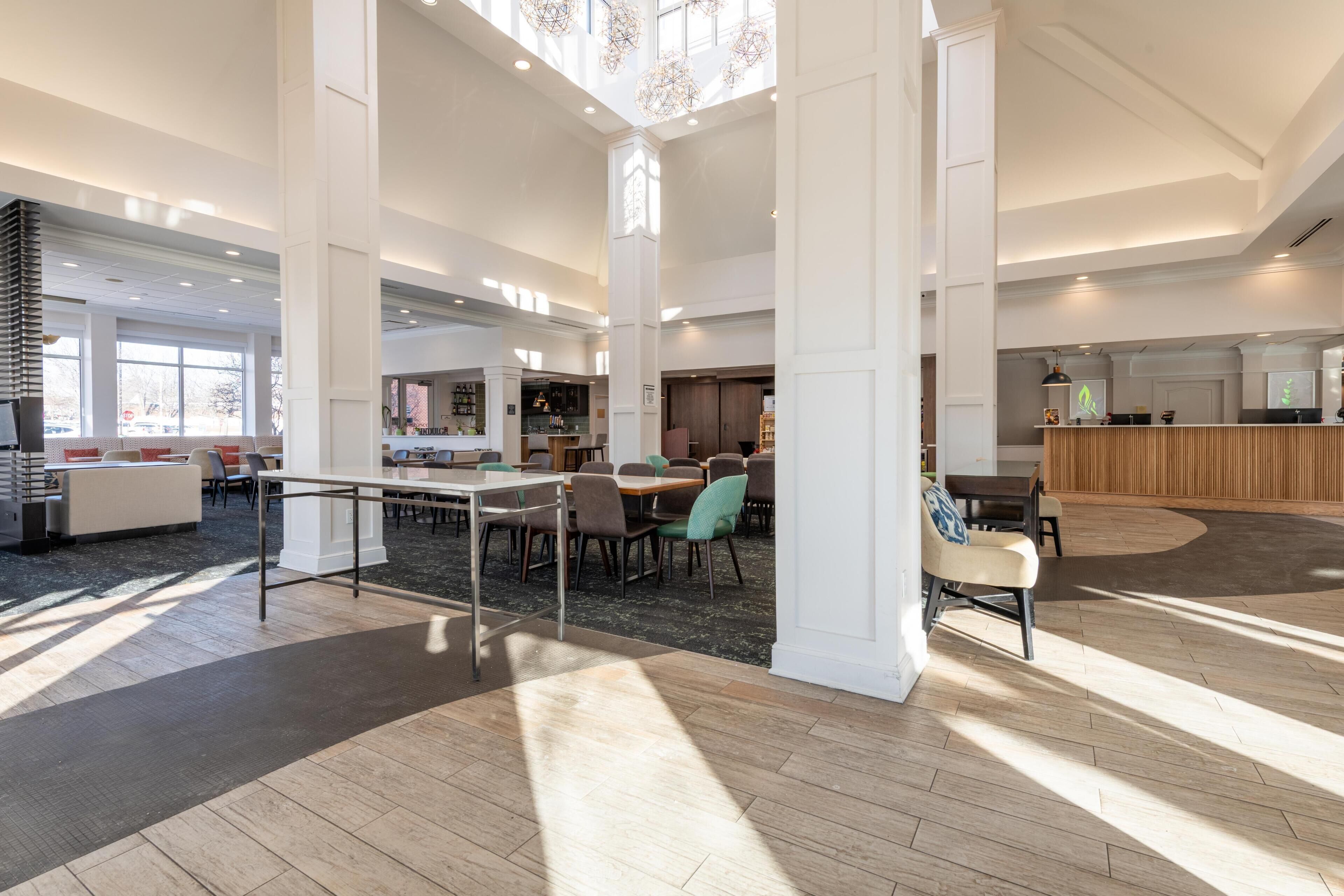 A spacious lobby with high ceilings, large windows, and a central seating area with tables and chairs.