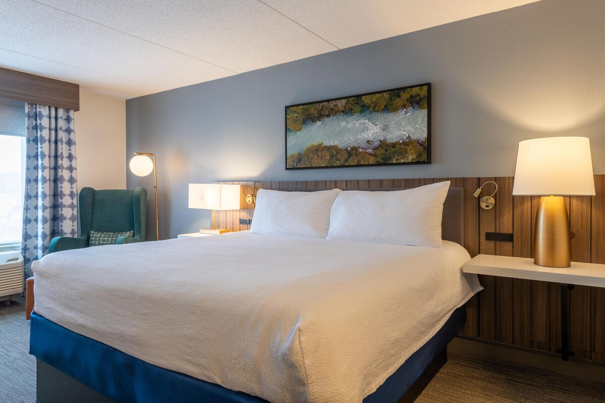 A hotel room with a large bed, two bedside lamps, a painting on the wall, and a chair.