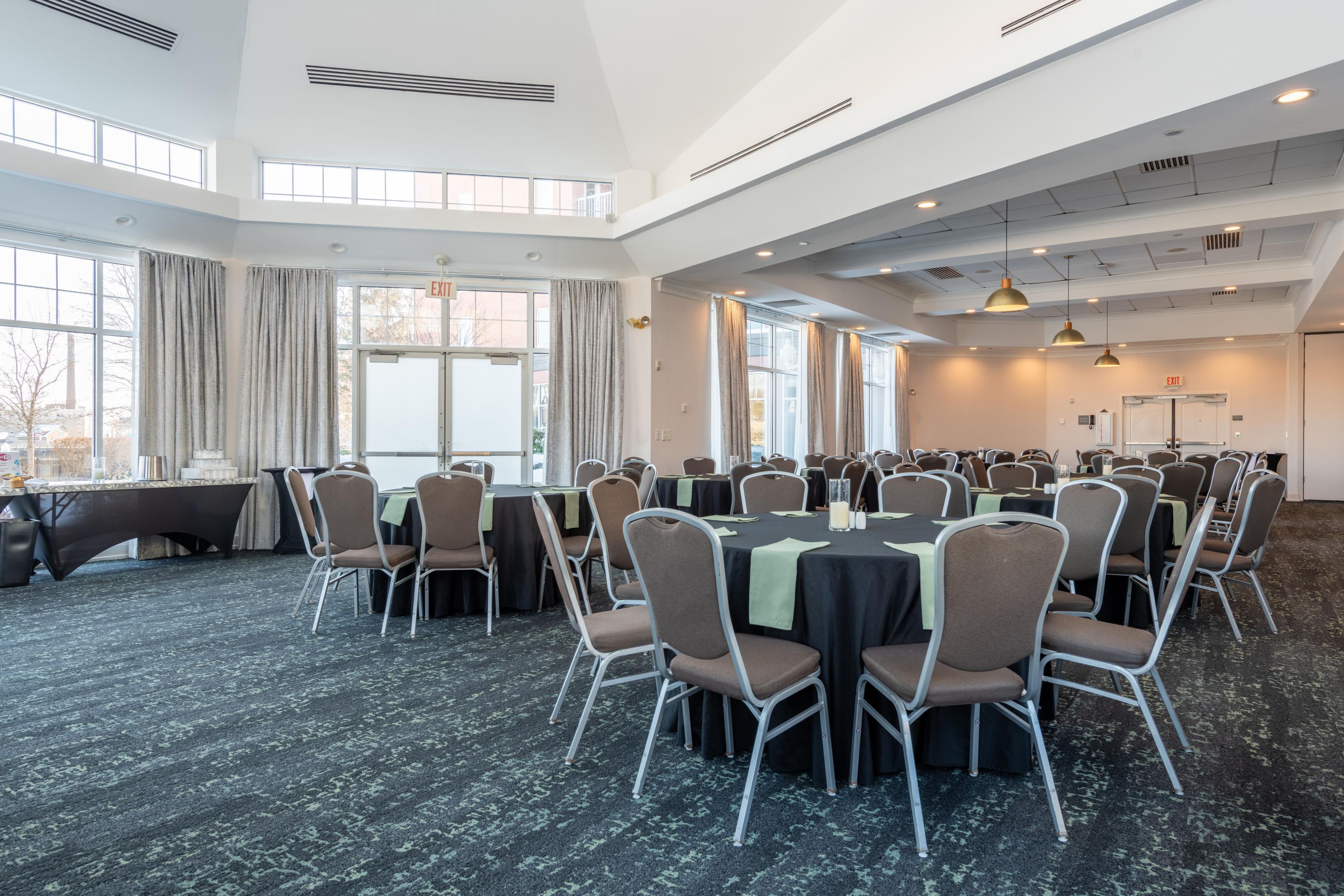 A conference room with multiple tables and chairs, large windows, and a high ceiling with ventilation.