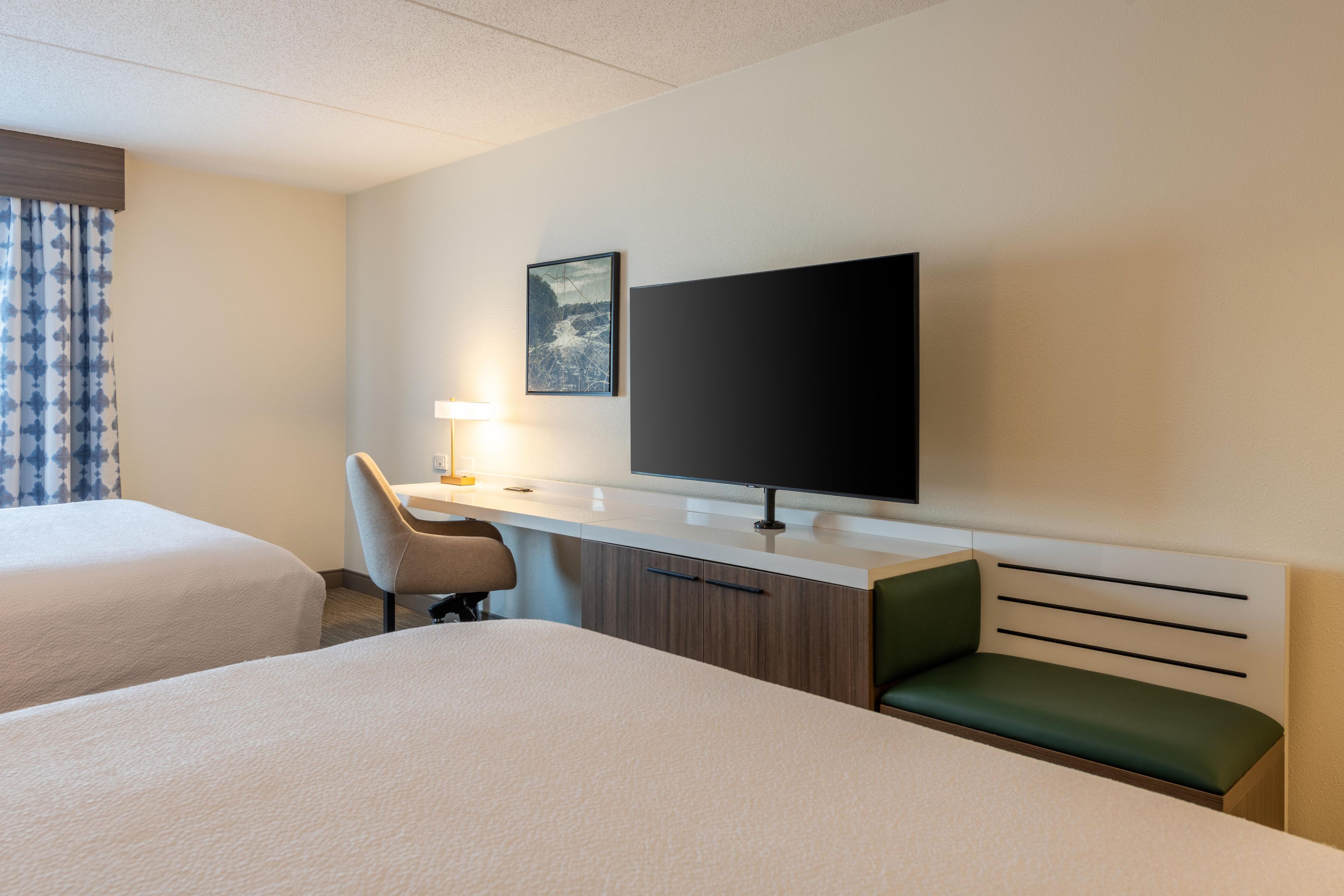 A hotel room with two beds, a desk with a chair, a flat-screen TV, a lamp, and a framed picture on the wall.