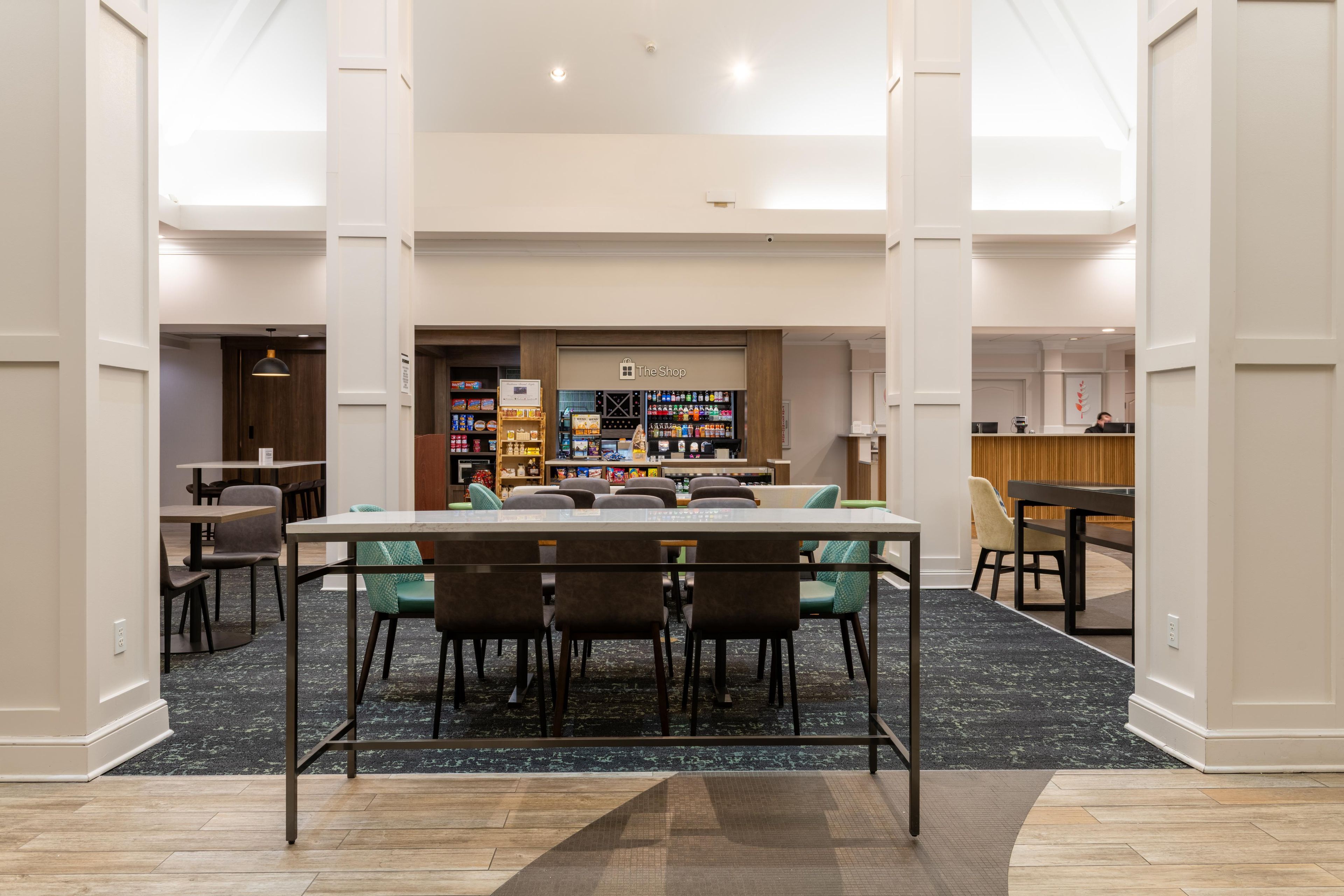 A hotel lobby with a reception desk, a seating area with a table and chairs, and a shop in the background.
