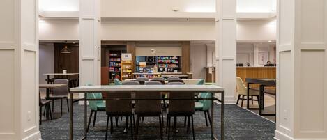 A hotel lobby with a reception desk, a seating area with a table and chairs, and a shop in the background.