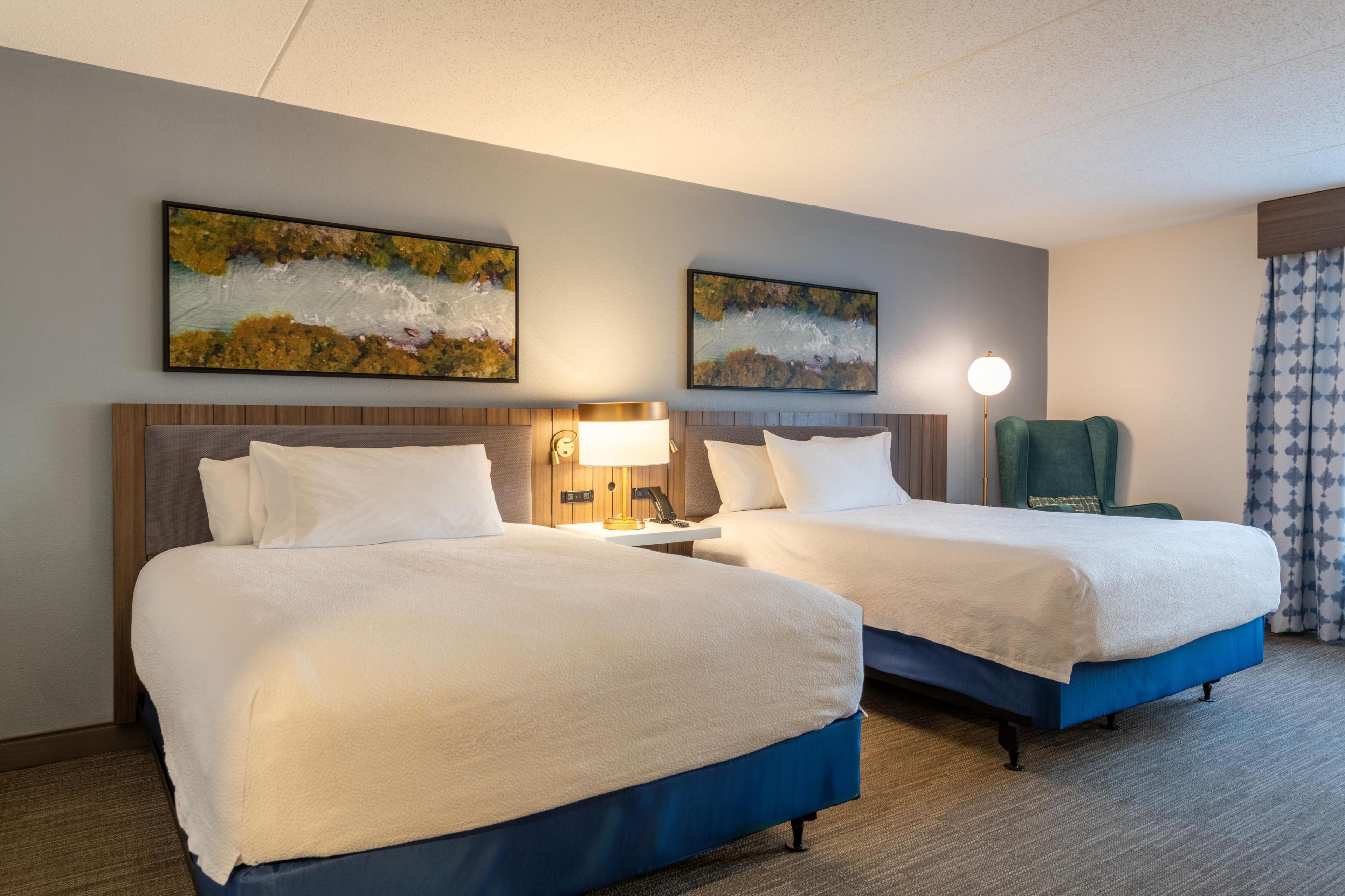 A hotel room with two beds, a nightstand with a lamp, a chair, and two wall-mounted paintings.