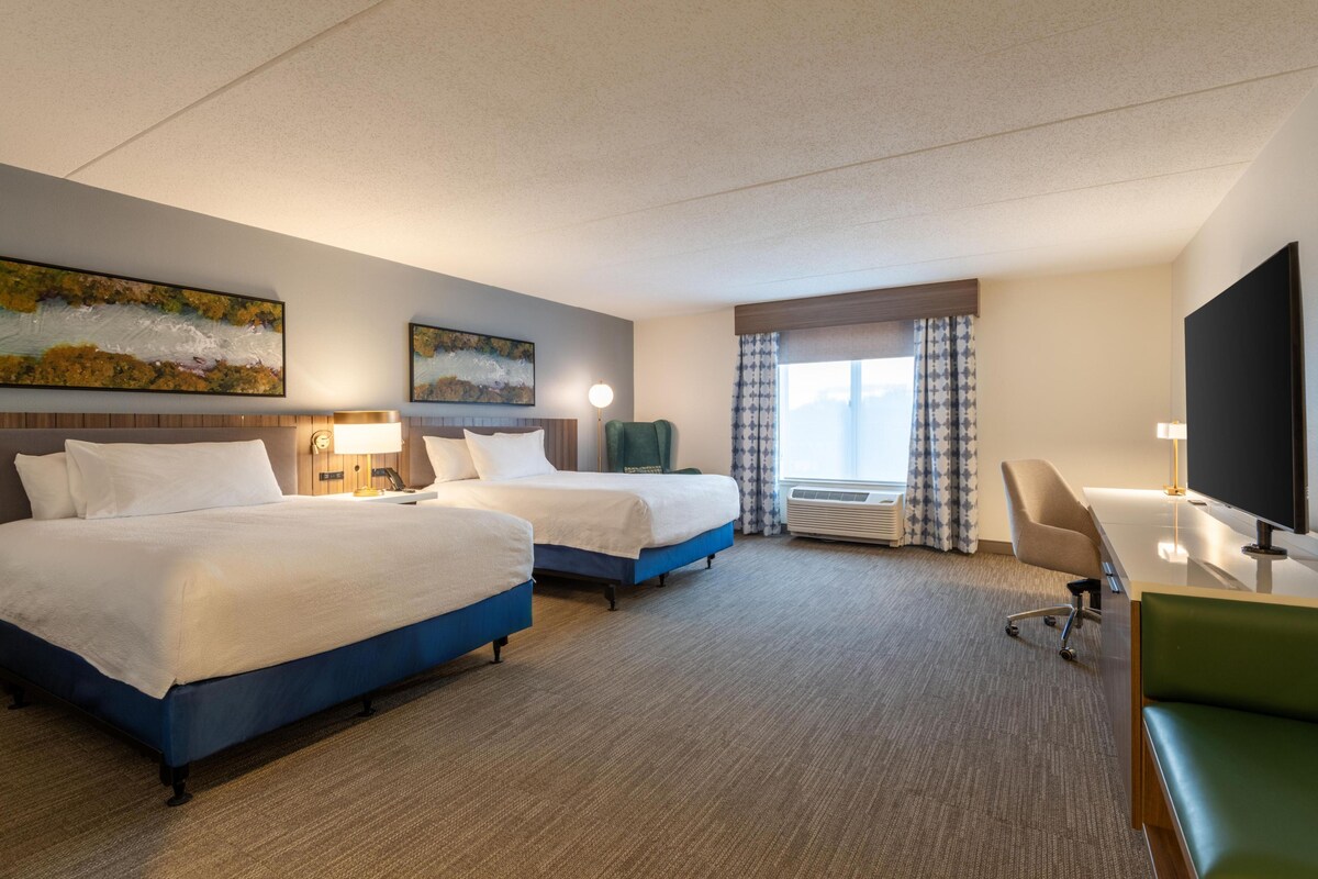 A hotel room with two beds, a desk, a chair, a TV, and a window with curtains.