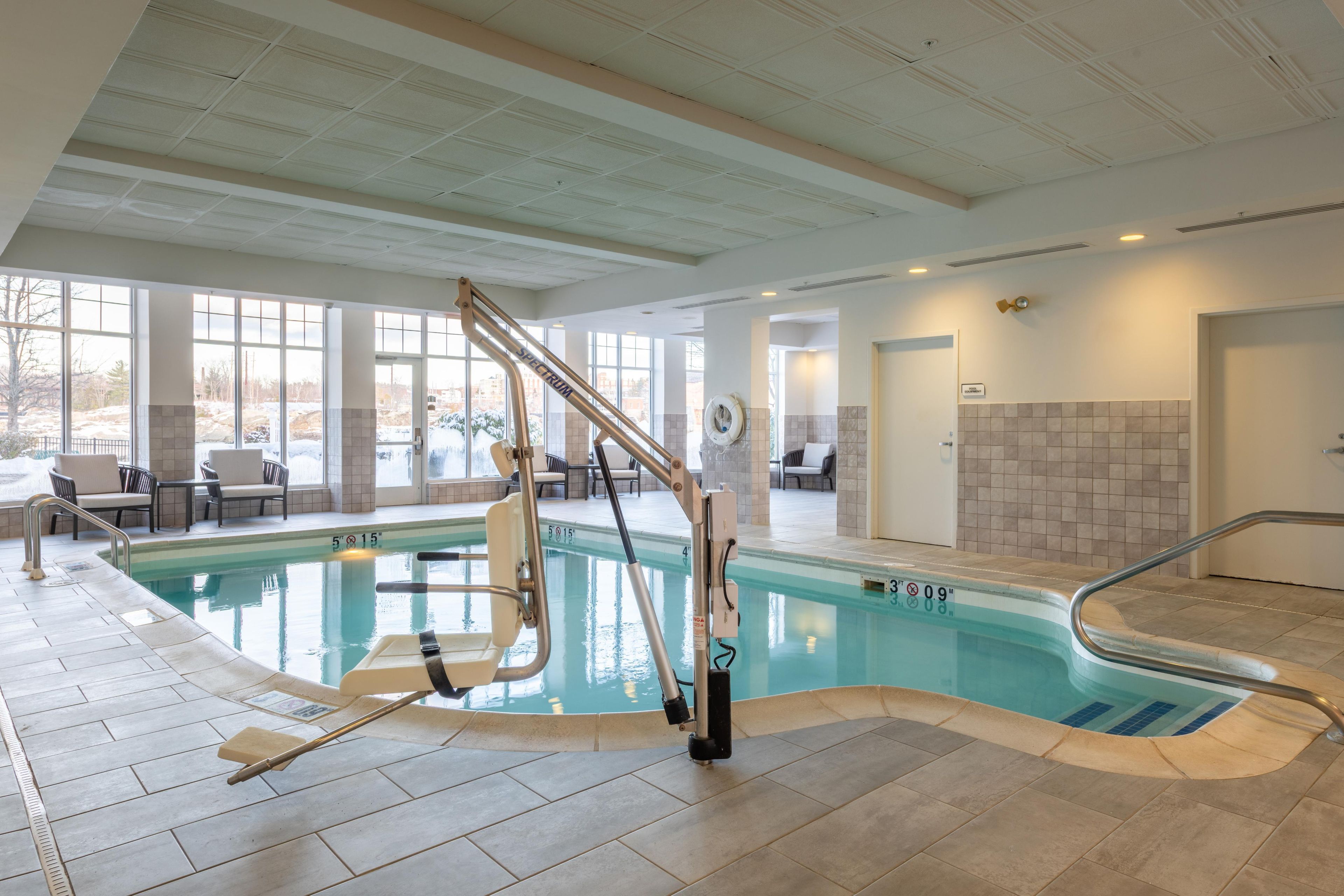 En indendørs swimmingpool med elevationssæde, flisebeklædte vægge og udsigt til det fri.