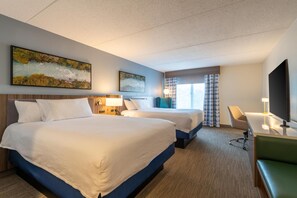 A hotel room with two beds, a desk, a chair, a TV, and artwork on the walls.
