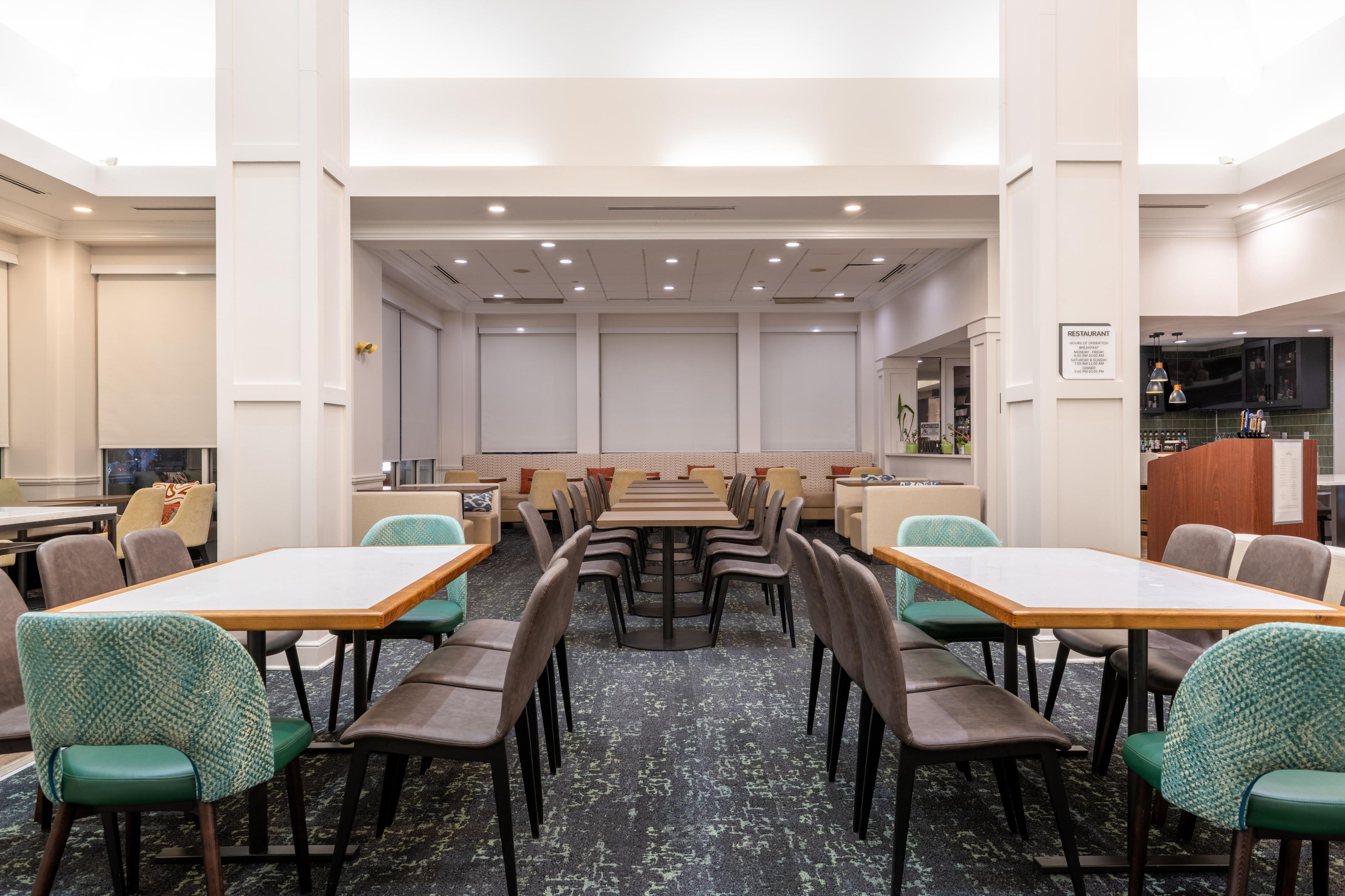 A hotel dining area with multiple tables and chairs, a kitchen area, and a seating area with sofas.