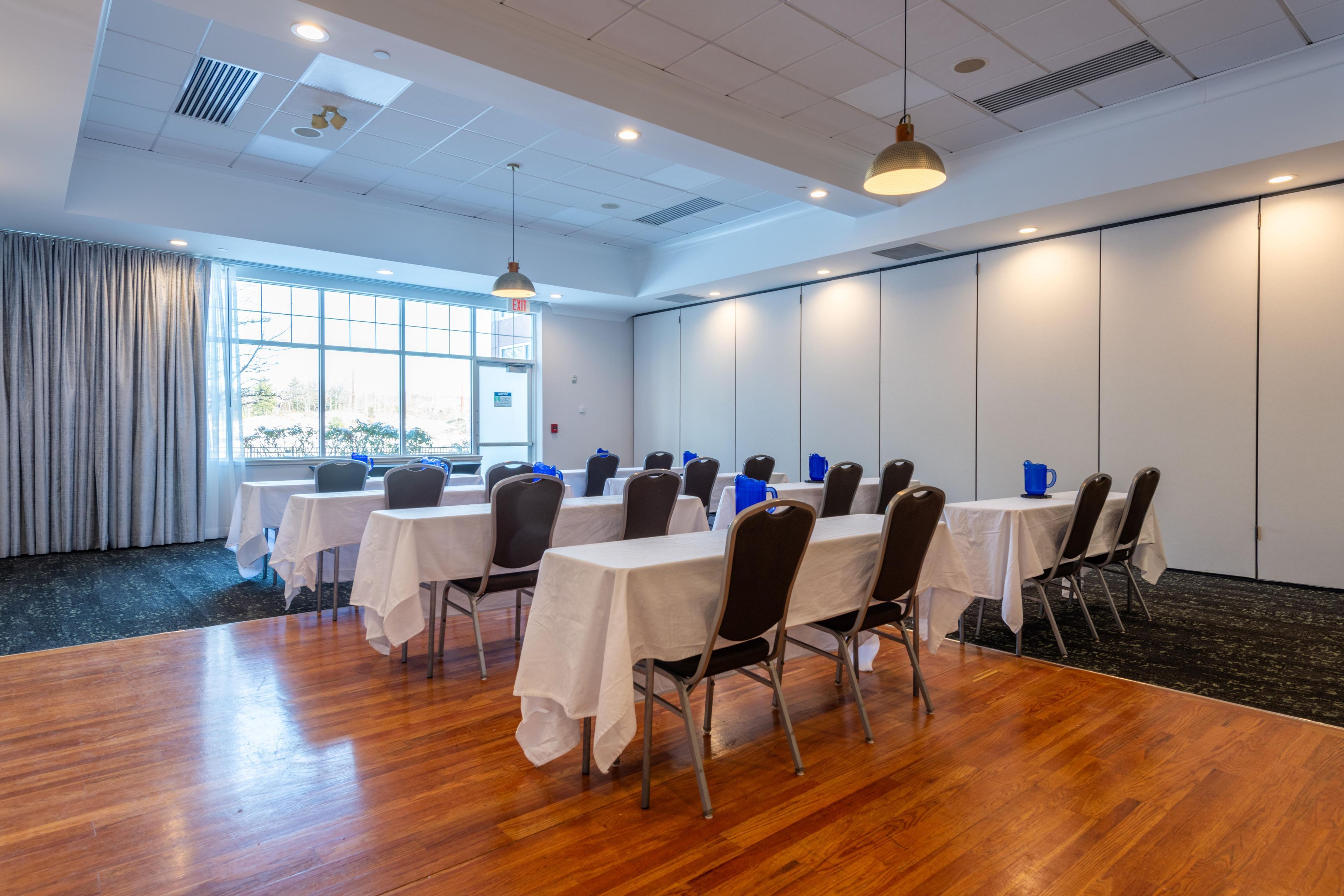 A conference room with white tables and chairs, wooden flooring, and large windows with curtains.