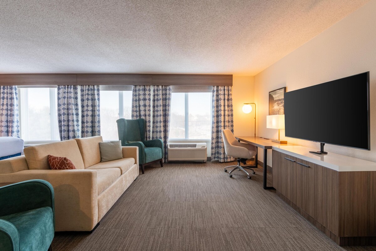 A hotel room with a sofa, a chair, a desk, a flat-screen TV, and a window with curtains.