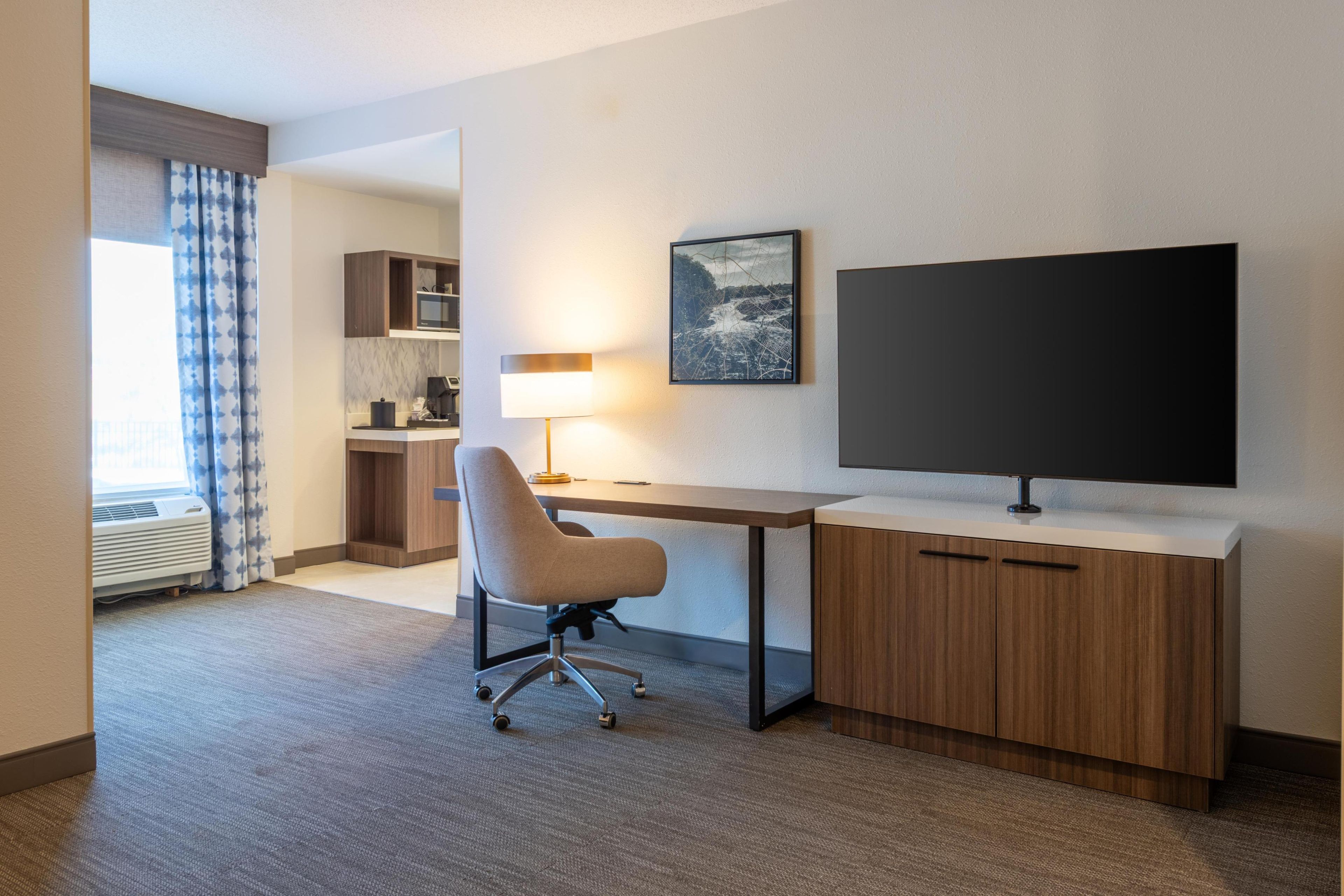 A hotel room with a desk, chair, TV, and kitchenette.