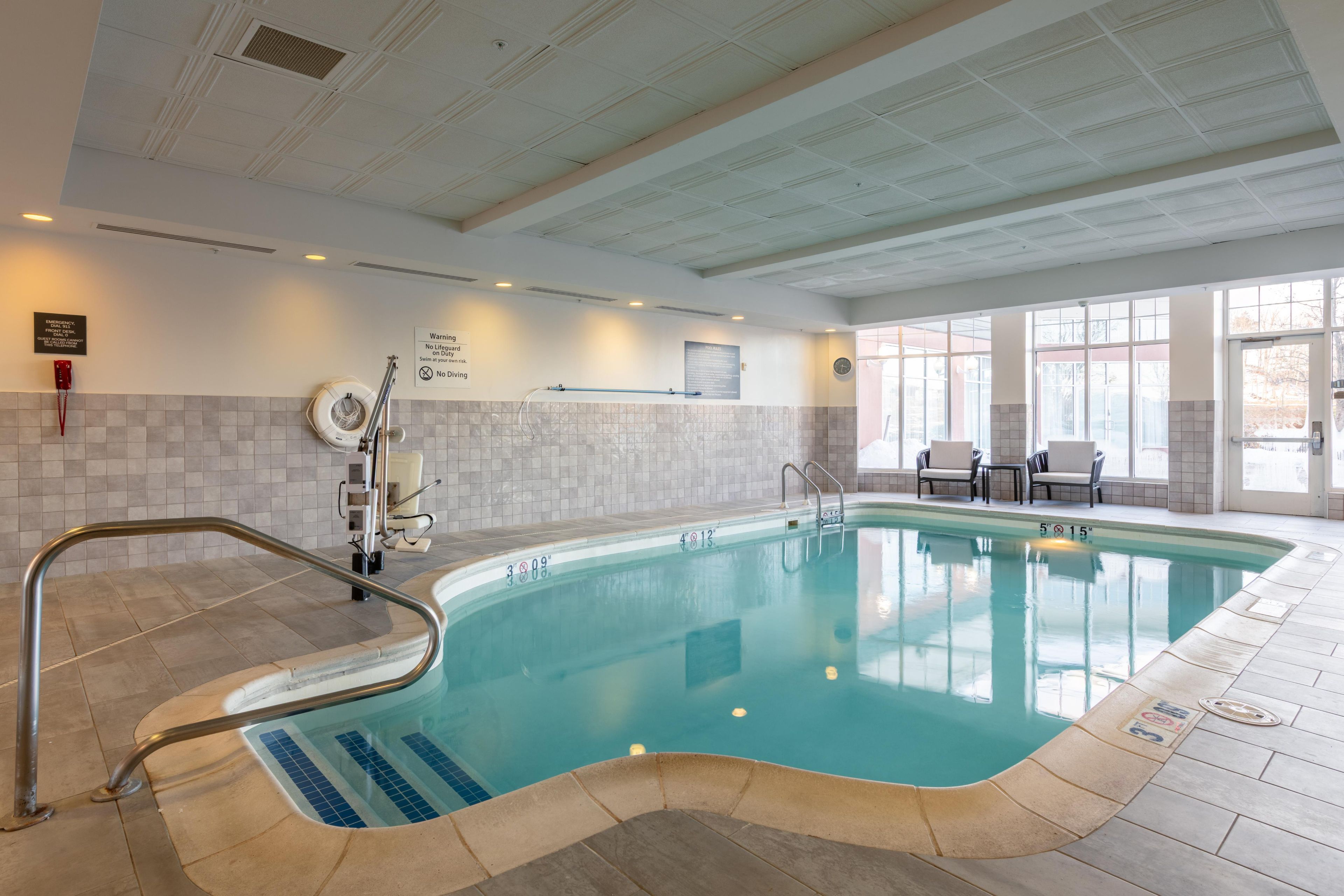 An indoor swimming pool with clear water, a lifeguard chair, and a seating area with chairs.