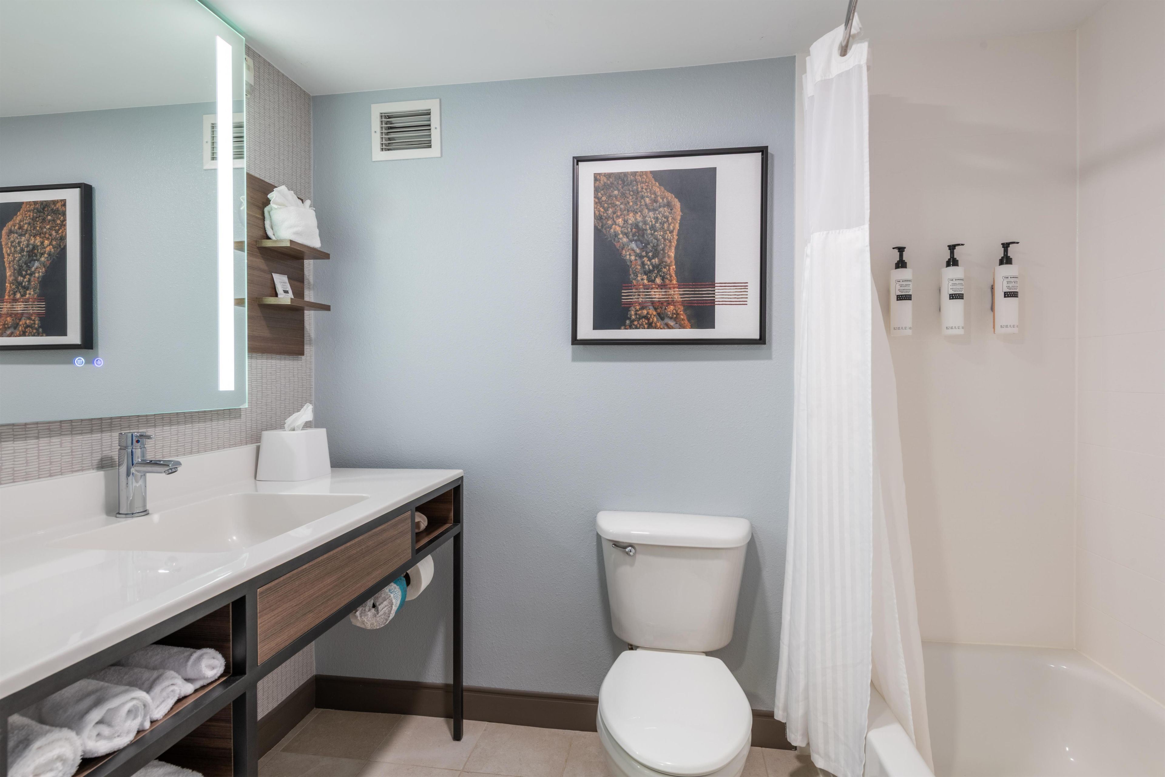 A bathroom with a white sink, toilet, and bathtub. There are towels on a shelf and a framed picture on the wall.