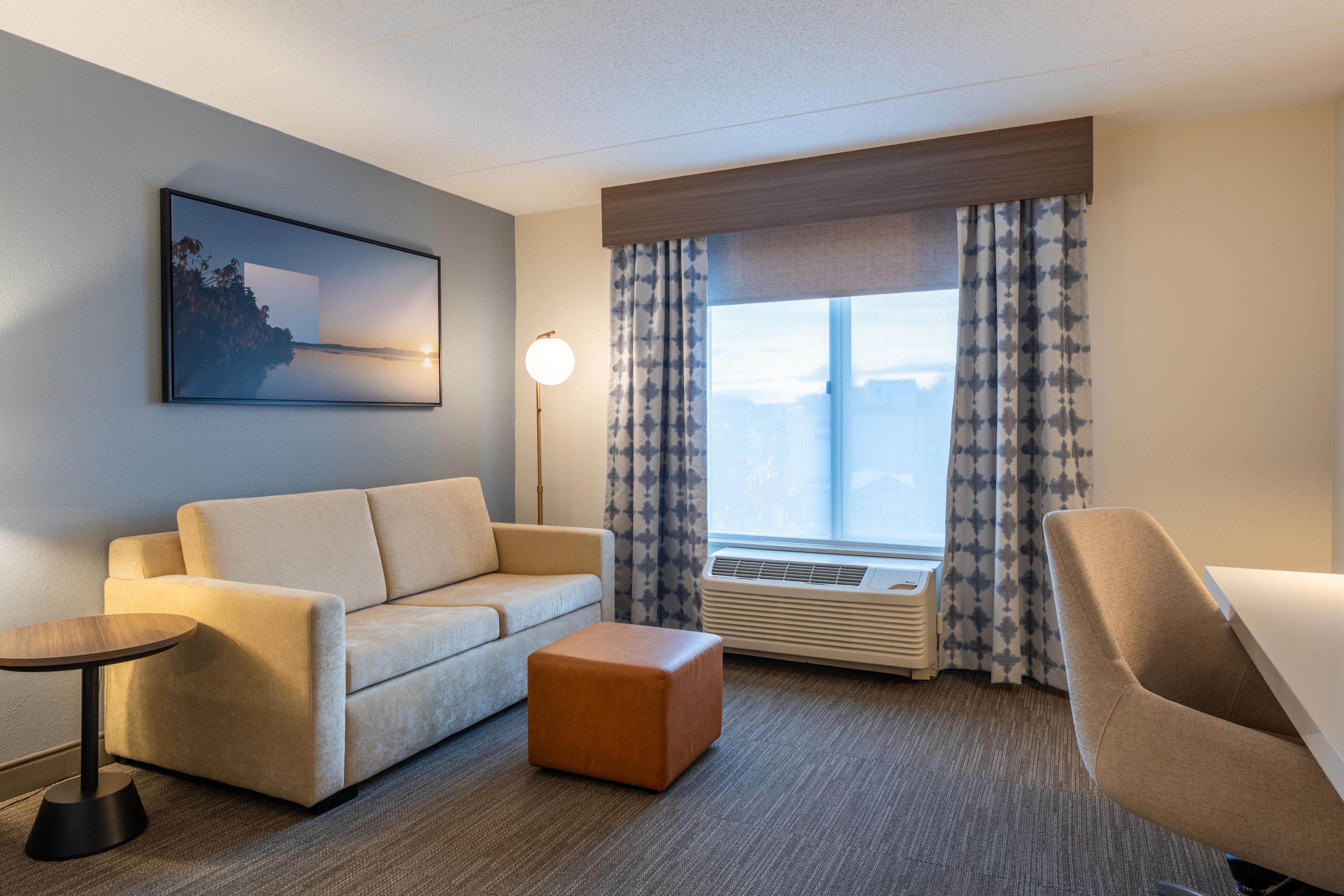 Habitación de hotel con sofá, reposapiés, una mesita, lámpara de pie y un amplio ventanal con cortinas.