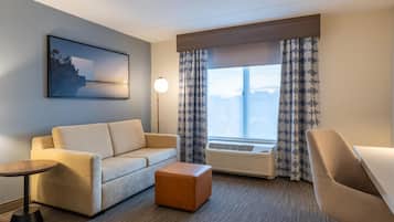 Habitación de hotel con sofá, reposapiés, una mesita, lámpara de pie y un amplio ventanal con cortinas.