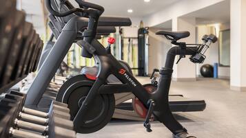 Una bicicleta estática en un gimnasio con el logotipo de Peloton, una caminadora y equipo de ejercicio al fondo.