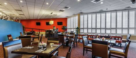 A spacious dining area with multiple tables and chairs, large windows with blinds, and a red accent wall.