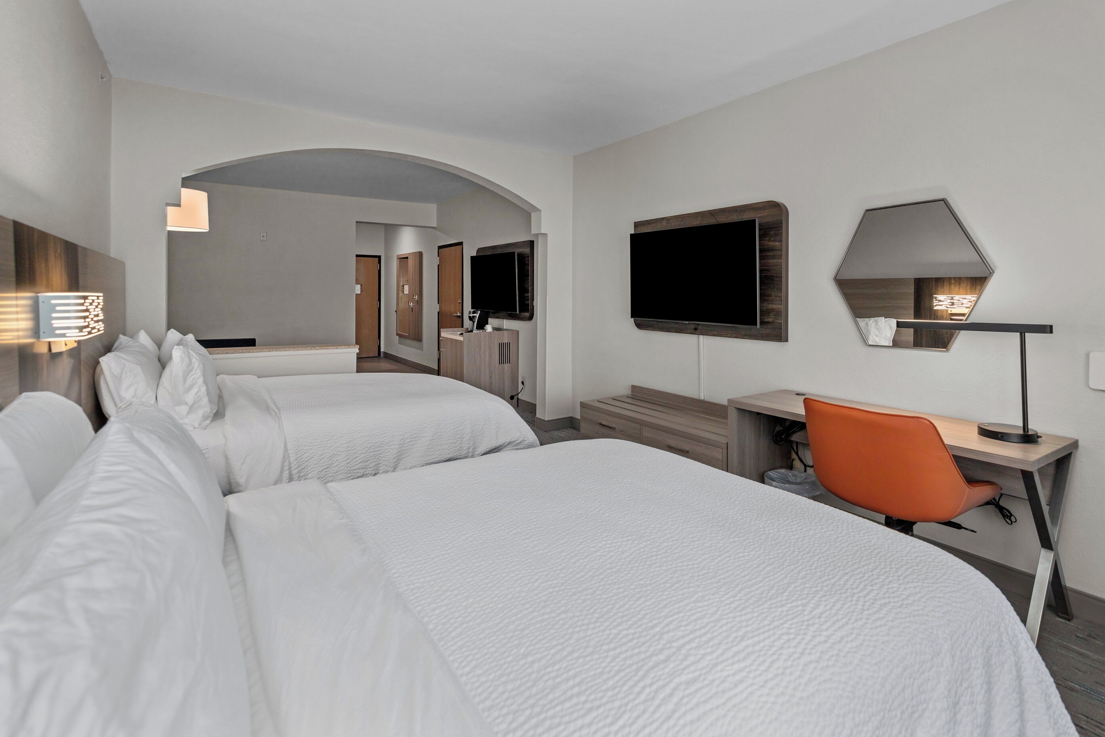 A hotel room with two beds, a desk, an orange chair, a TV, and a mirror.