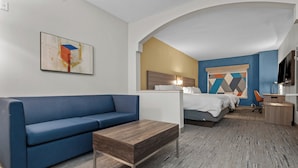 A hotel room with a blue sofa, a wooden coffee table, a bed with a white comforter, a desk with a lamp, and a television. - Holiday Inn Express Crockett by IHG (Crockett)