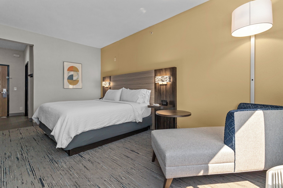A modern hotel room with a large bed, a small round table, a chair, and a wall-mounted lamp.