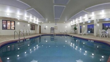 Una piscina interior con agua azul cristalina, rodeada de sillas y mesas blancas.