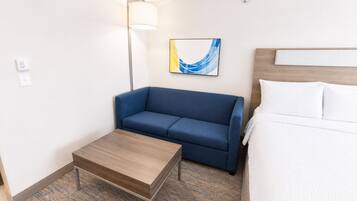 A hotel room with a blue sofa, a wooden coffee table, a bed with white bedding, a lamp, and a wall art piece.