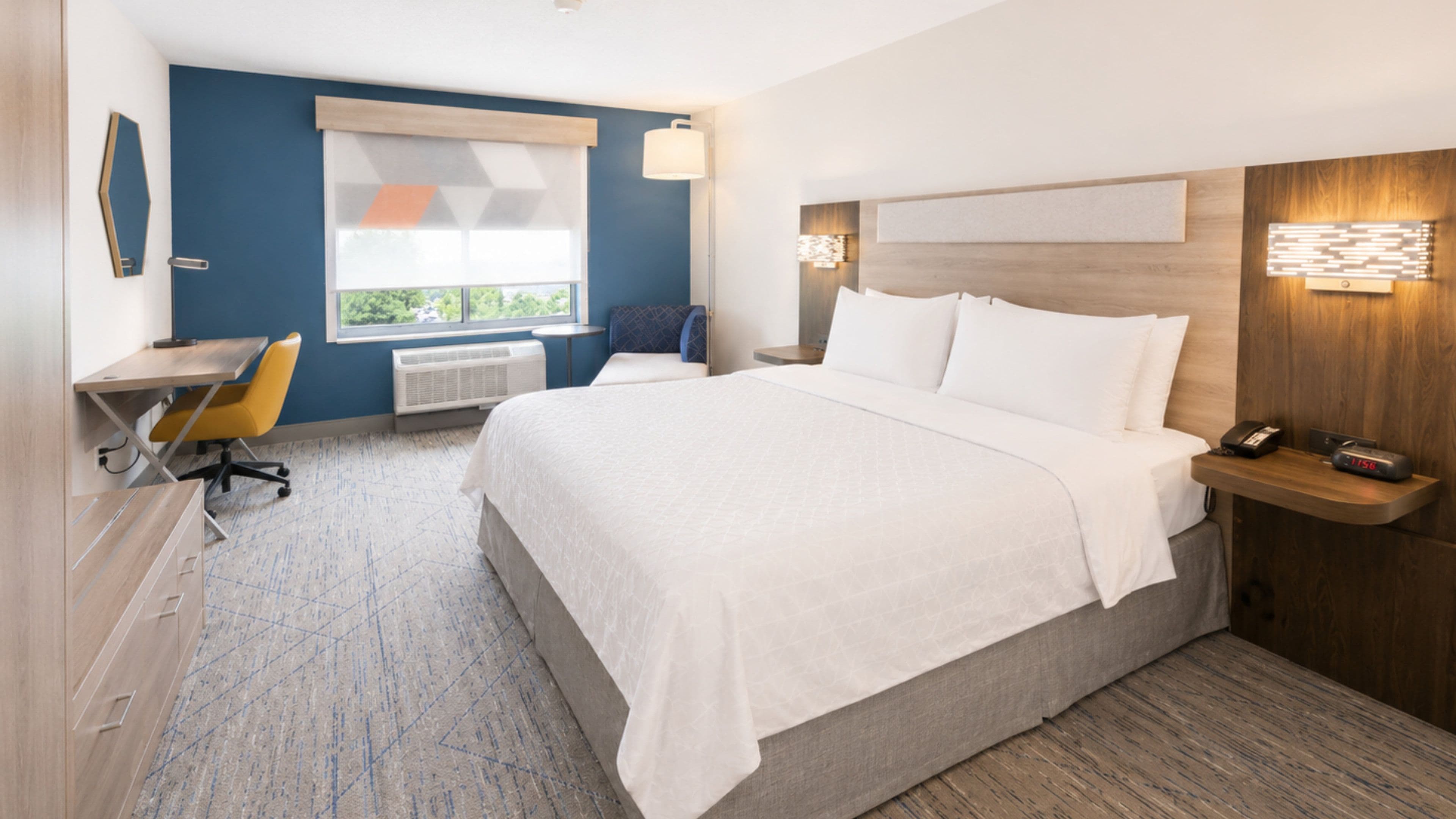 A hotel room with a bed, desk, chair, bedside table, lamp, and a window with blinds.
