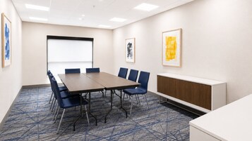 Un salón de conferencias con una gran mesa, sillas azules, una pantalla de proyector y obras de arte enmarcadas en las paredes.