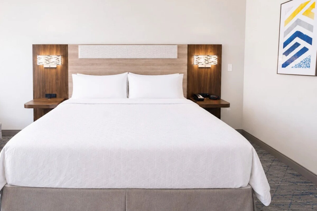 Een hotelkamer met een groot bed, twee nachtkastjes, een lamp en een kunstwerk aan de muur.
