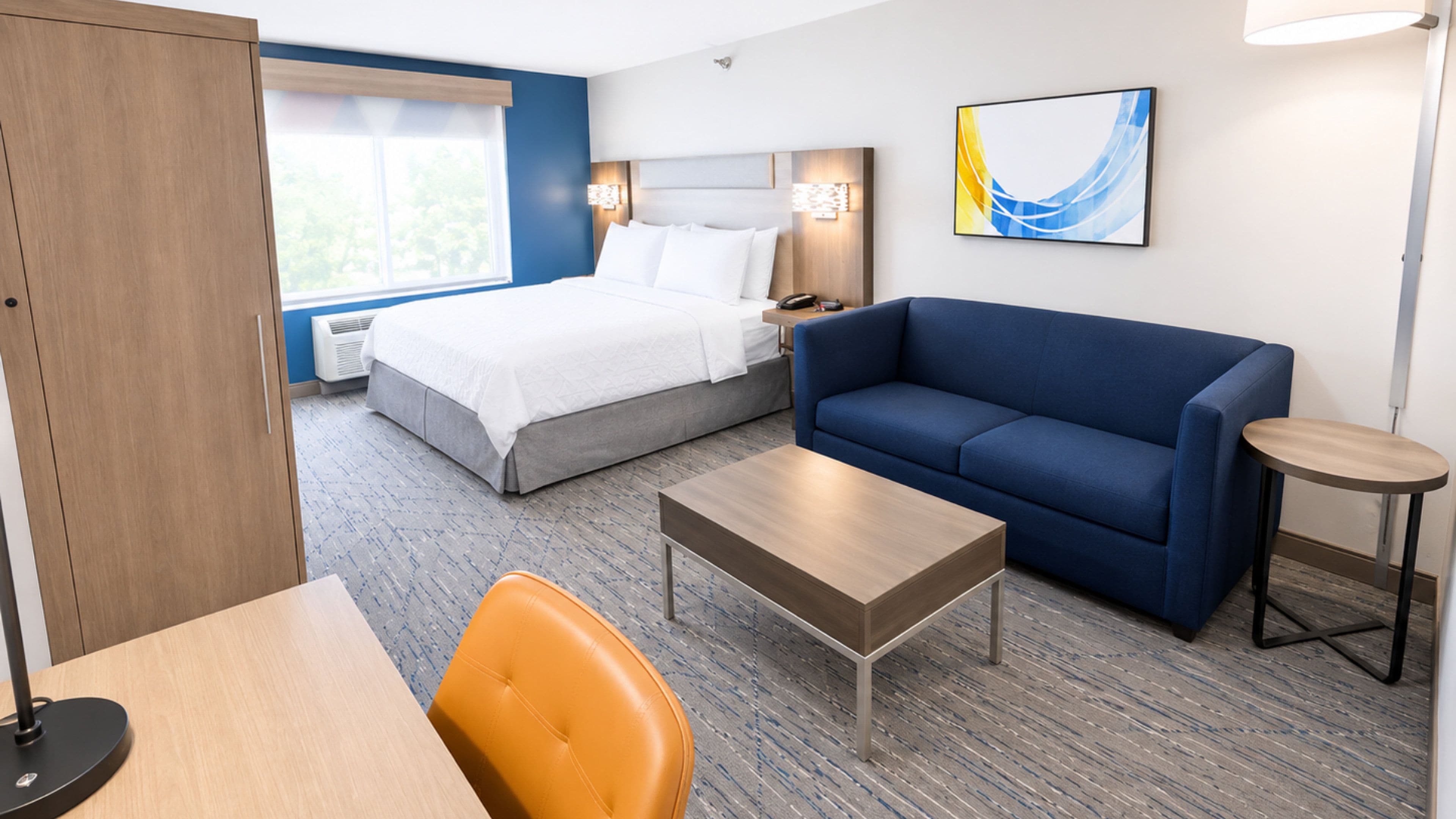 A hotel room with a bed, a sofa, a coffee table, a chair, a desk, a lamp, and a painting on the wall.