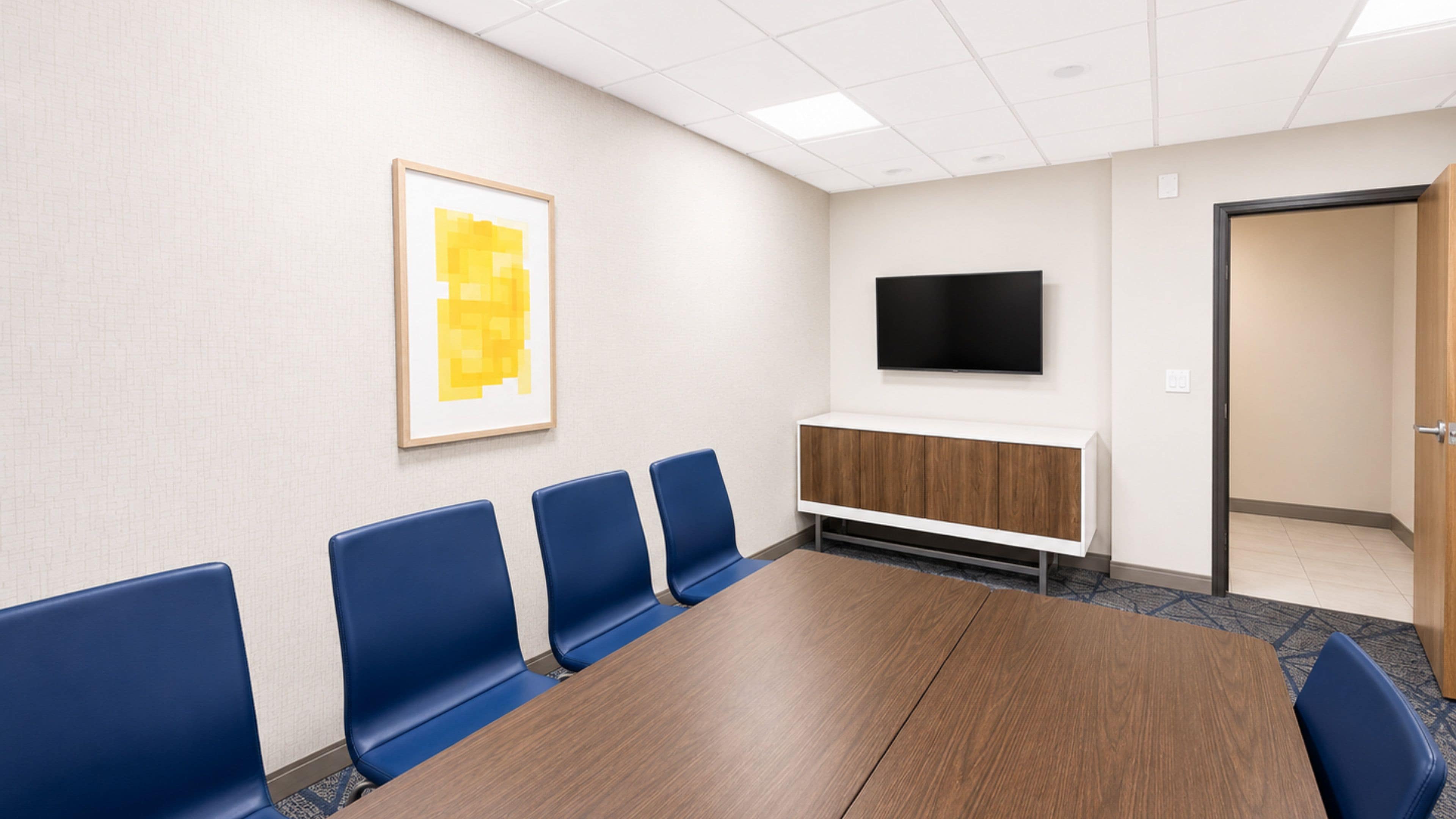 A conference room with a long table, blue chairs, a mounted TV, and a framed picture on the wall.