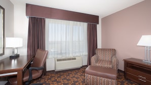 A hotel room with a desk, two chairs, a sofa, a lamp, and a window with curtains.