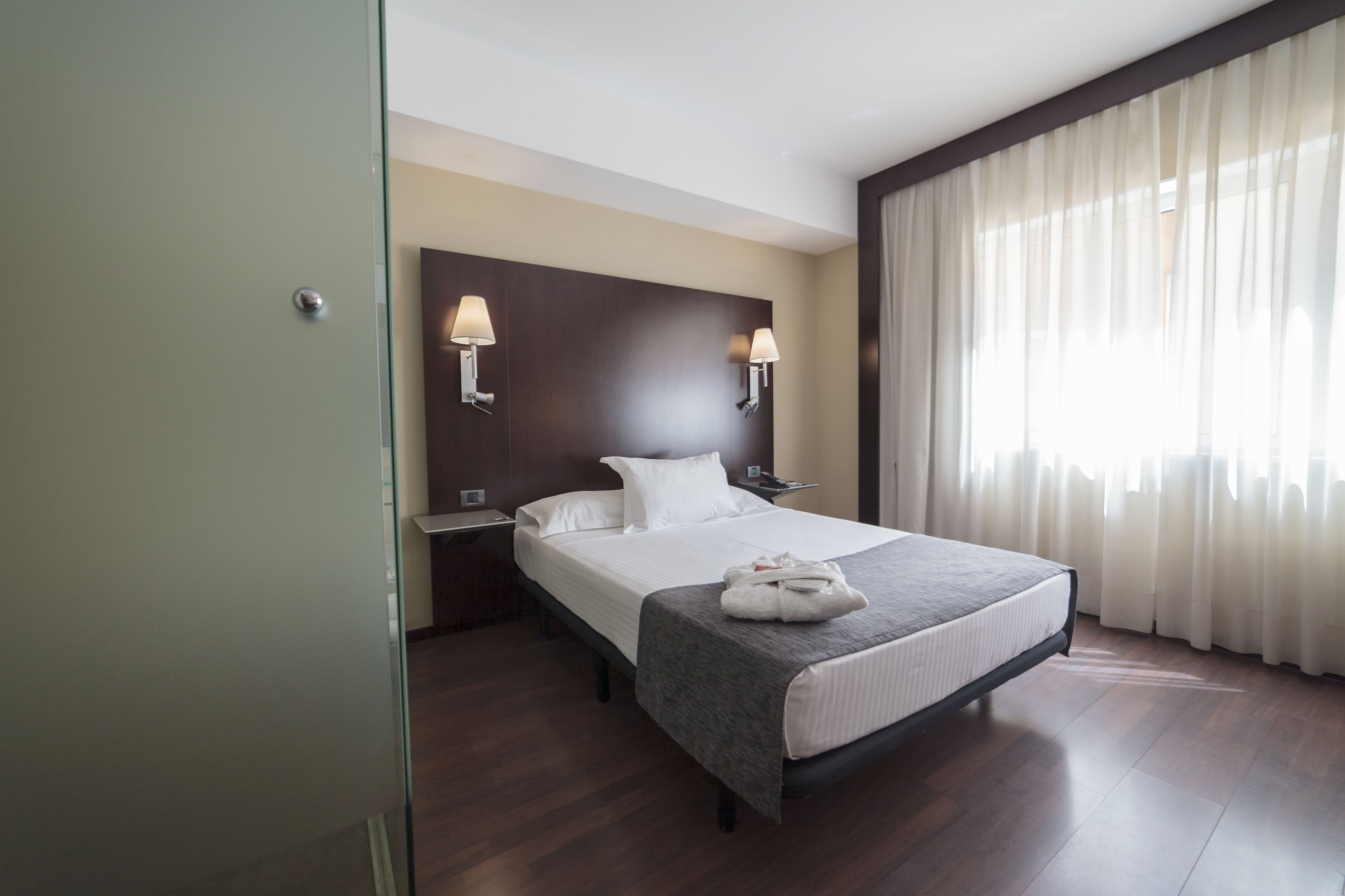 Una habitación de hotel con una cama grande, dos lámparas de noche, una mesita de noche y una ventana con cortinas.