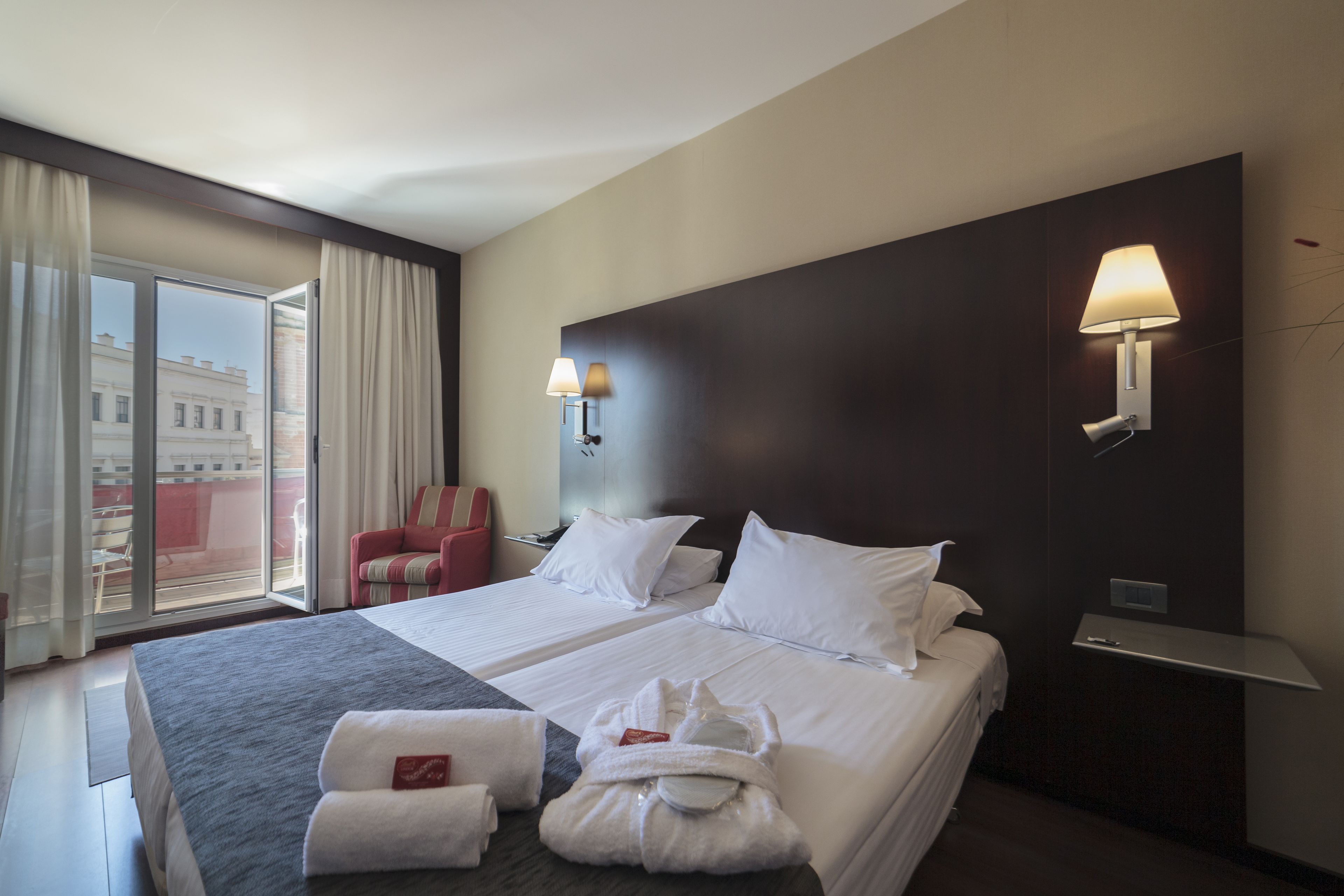 Une chambre d’hôtel avec un grand lit, deux lampes de chevet, un fauteuil rouge, une fenêtre avec des rideaux et une vue sur les bâtiments environnants.
