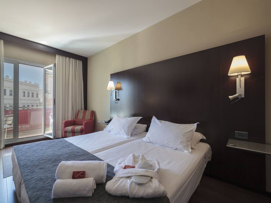 A hotel room with a large bed, two bedside lamps, a red chair, a window with curtains, and a view of buildings outside.