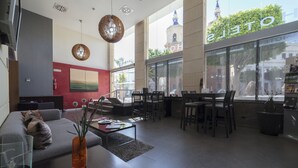 A modern hotel lobby with a sofa, dining area, and large windows offering views of a cityscape and a clock tower. - Hotel Salymar (San Fernando)