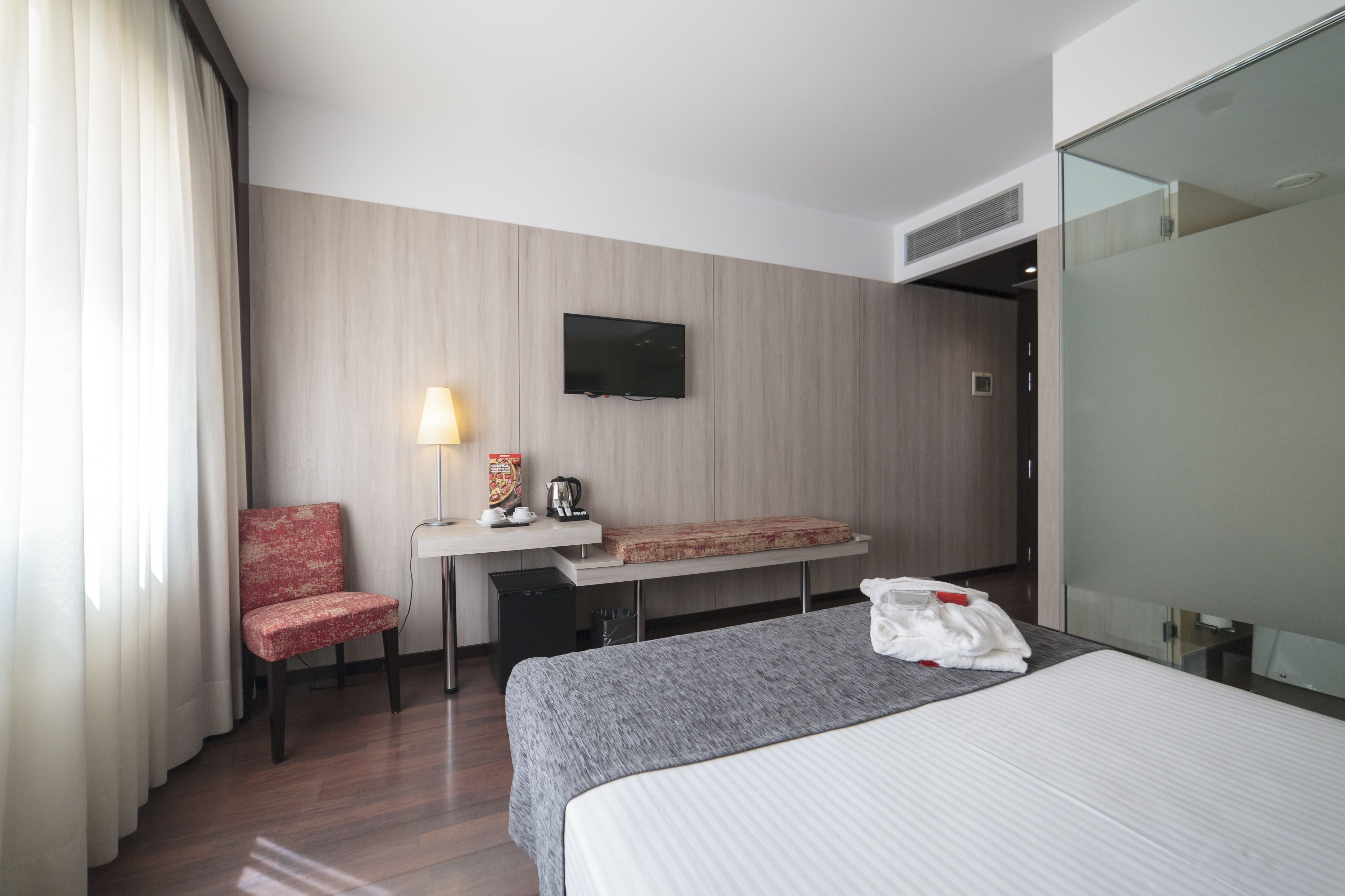 Une chambre d’hôtel comprenant un lit, un bureau avec une lampe, une chaise, un téléviseur fixé au mur et une cloison en verre.