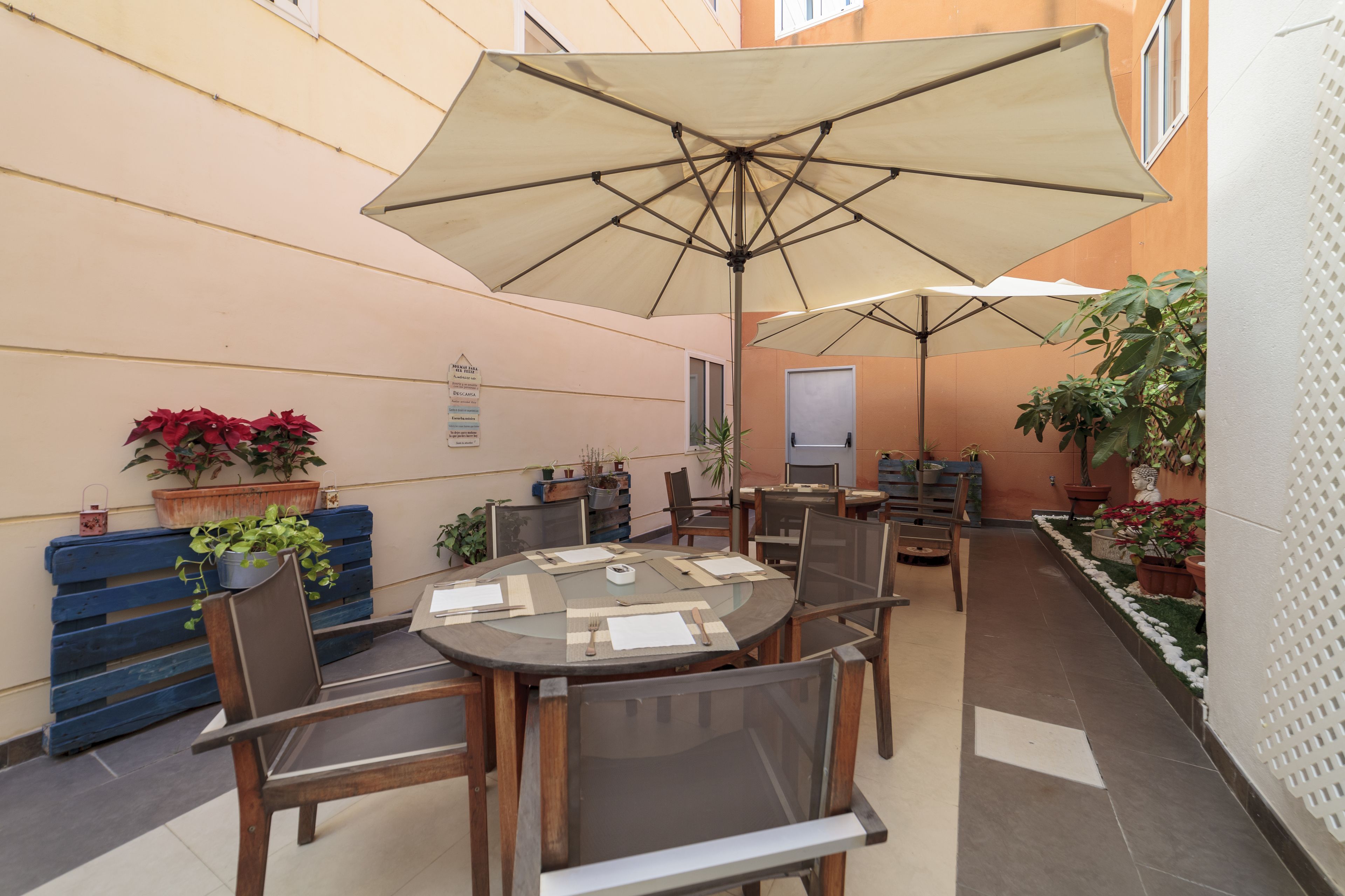 A patio area with a round table, chairs, and a large umbrella. There are potted plants and a blue bench along the wall.